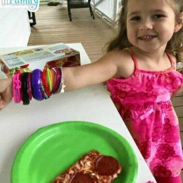 A little girl standing at a table with a piece of pizza in front of her.