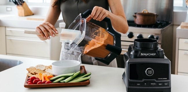  A woman pouring dip from a Ninja blender into a bowl with cut vegetables.