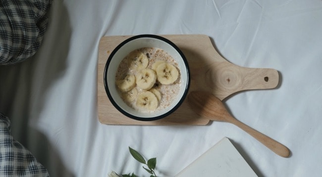 A white bowl of oatmeal and sliced bananas resting on a wooden cutting board.