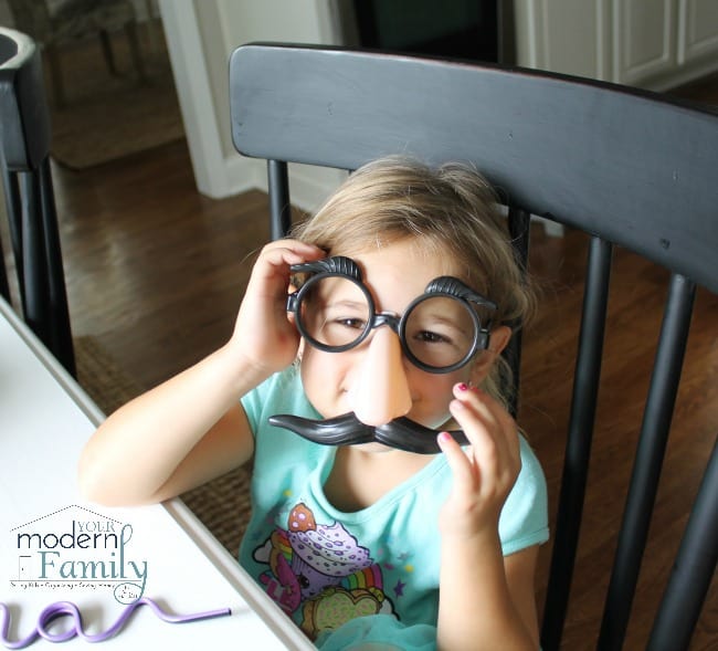 A girl sitting at a table wearing fake glasses , nose and mustache.