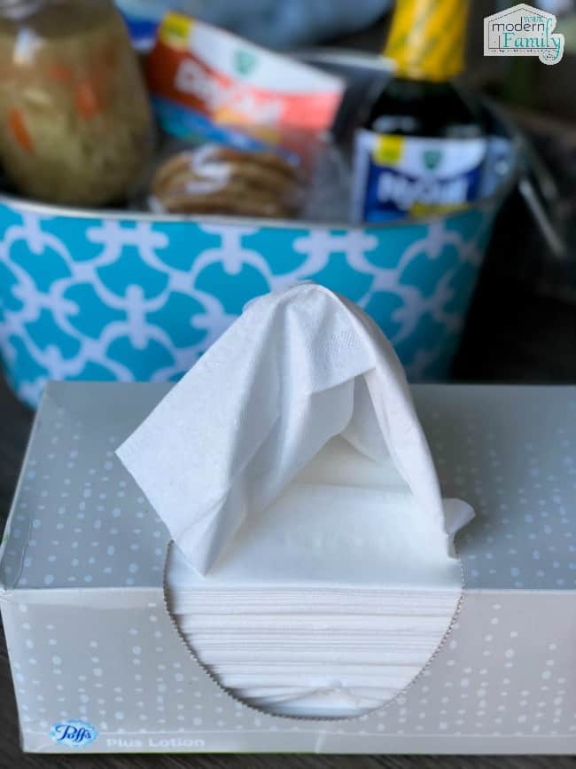 A close up of a tissue box and a blue basket of items behind it.