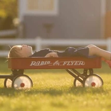 A young boy lying on a red wagon looking up to the sky.
