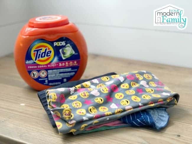 A container of Tide Pods detergent beside colorful children\'s clothes neatly folded.