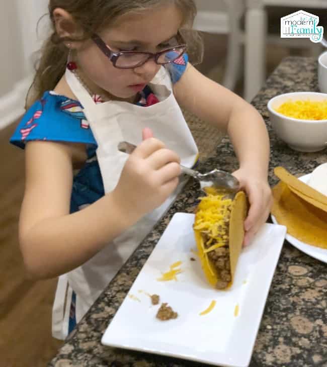 A little girl wearing a white apron sitting at a table putting cheese on a taco.