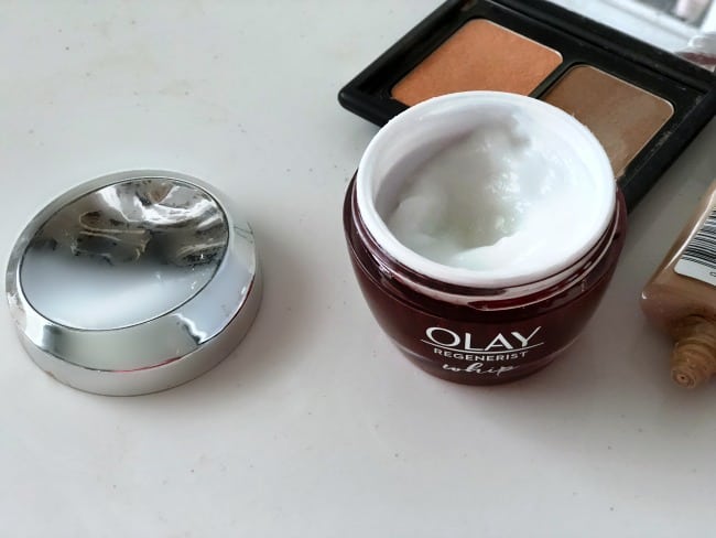 An open jar of Olay Regenerist Whip with make up behind it.