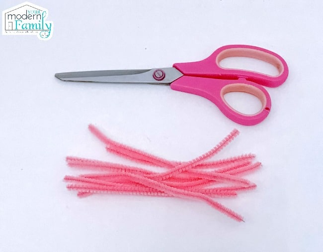 A pair of scissors lying beside pink pipe cleaners.