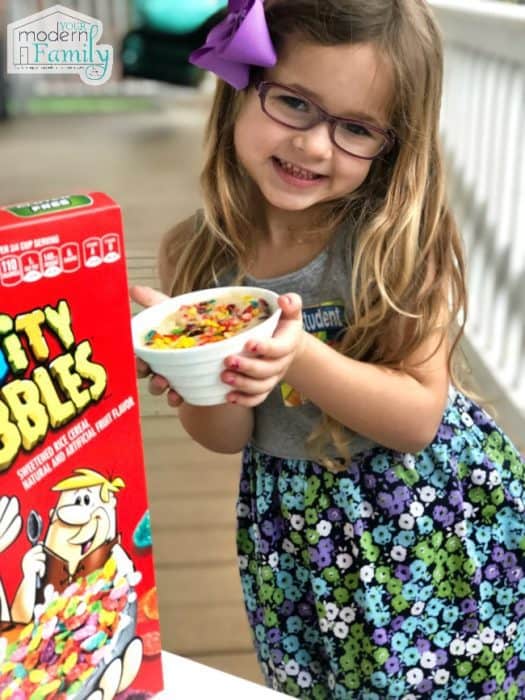A little girl holding a microwave cake in a bowl with a box of Fruity Pebbles beside her.