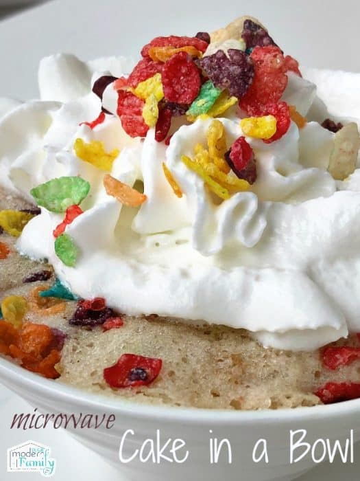 A close up of Microwave cake in a bowl with whipped cream and Fruity Pebbles sprinkled on top.