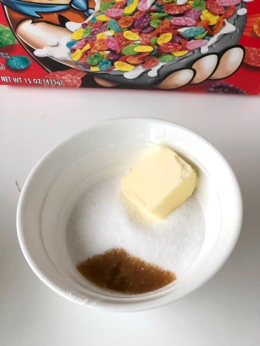 A white bowl sitting on  a table with a pat of butter and brown sugar in it with a box of Fruity Pebbles behind it.