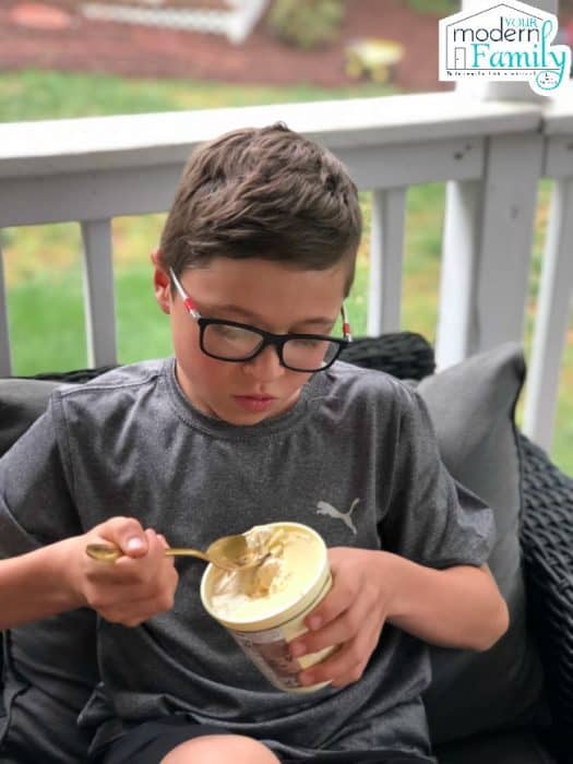 A boy sitting on a porch eating a container of ice cream with a golden spoon.
