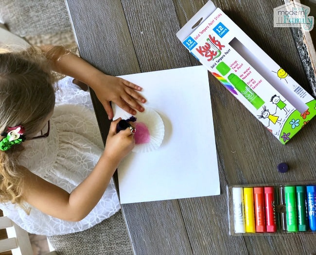 A little girl painting at a table with paint sticks.