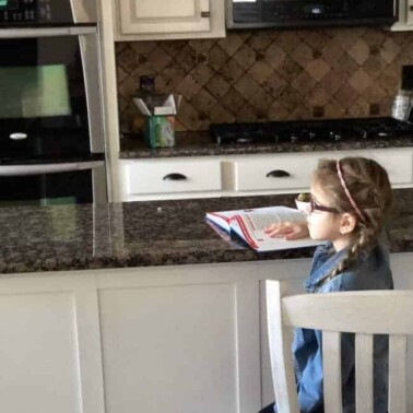 A girl sitting on at a kitchen counter with an open book in front of her.