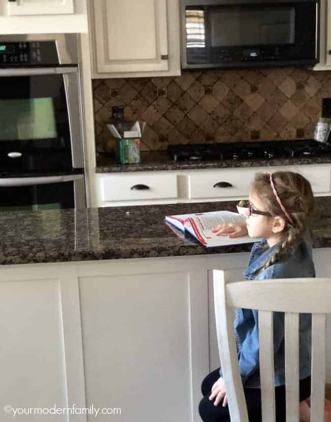 A  girl sitting on at a  kitchen counter with an open book in front  of her.