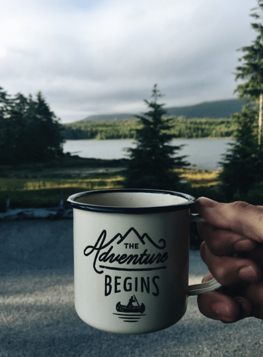 A person holding a cup of coffee with a view of trees and a lake in the background.