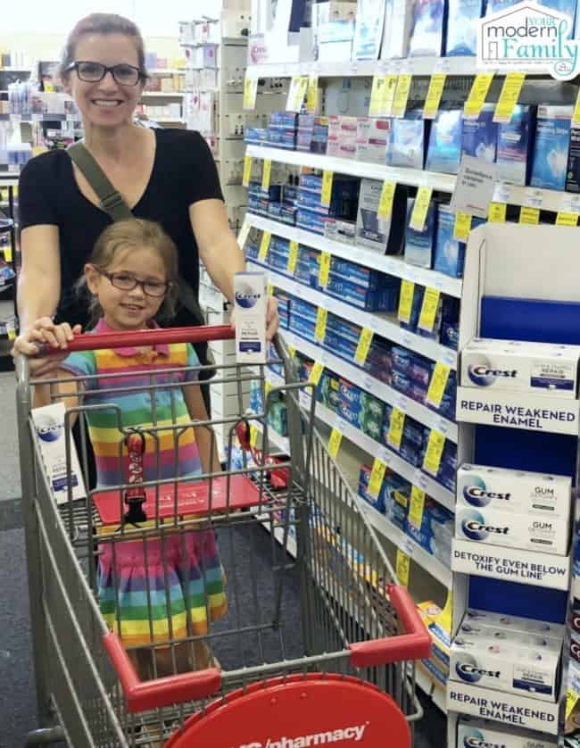 A woman and a child pushing a shopping cart while holding a box of Crest.