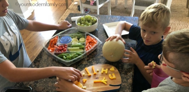 A group of boys standing at a table with a tray of fruit and vegetables and a platter of cheese cut into letters and shapes.