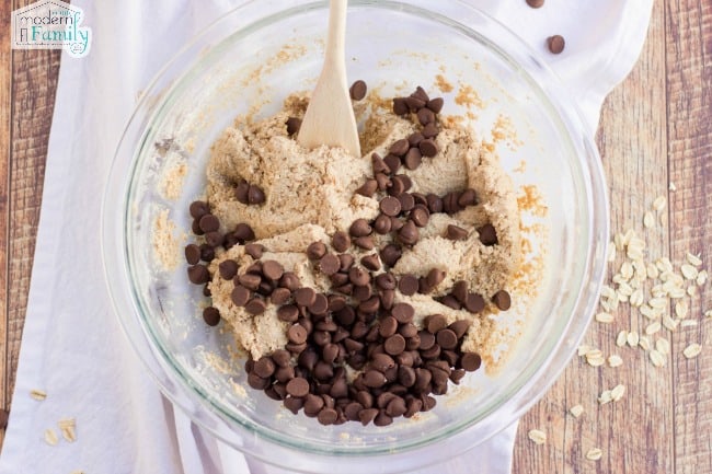 A clear glass bowl with chocolate chip cookie ingredients and a wooden spoon.