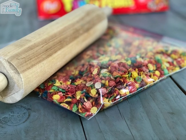  A clear baggie of Fruity Pebbles resting on a table with a wooden rolling pin beside it.