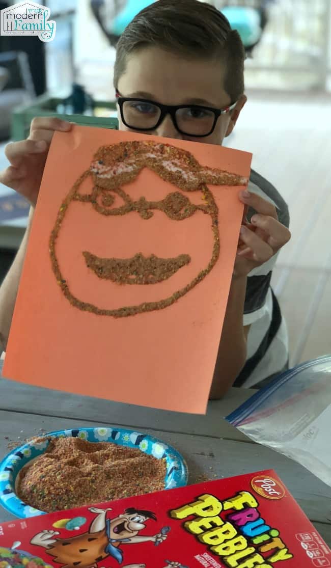 A child holding a piece of paper with a completed crushed cereal face design.