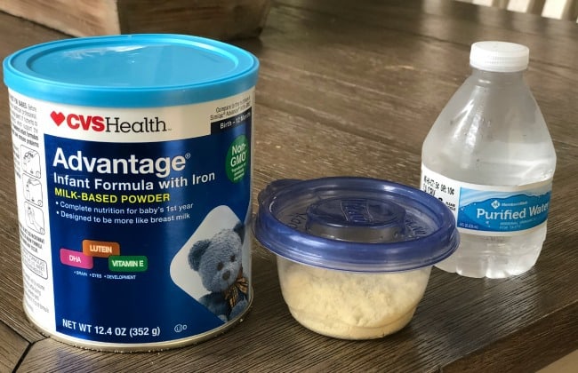 A can of baby formula with a clear plastic container with a blue lid beside it with a small bottle of water.