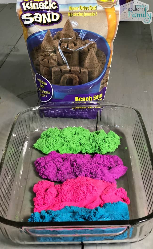 A glass container of colorful Kinetic Sand with a bag of brown sand in a bag behind it.