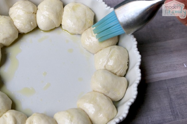 Biscuit balls in a round pan with a blue utensil adding butter.
