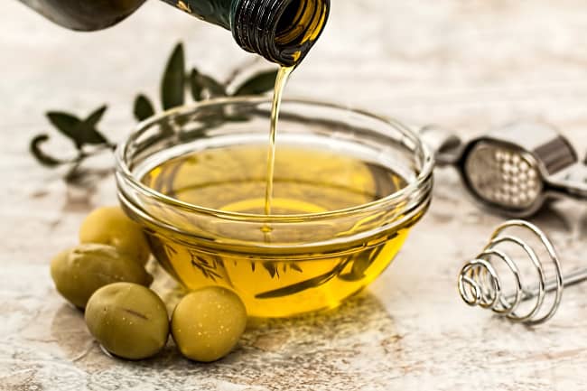  A picture of olive oil being poured from the bottle into a glass bowl with olives resting around the outside of the bowl.