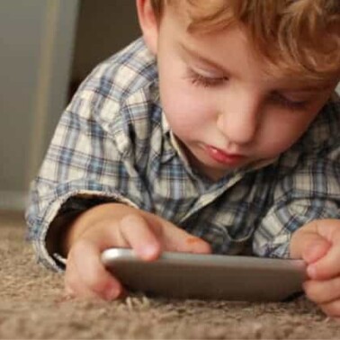 A young boy lying on a carpeted floor looking at a cell phone.