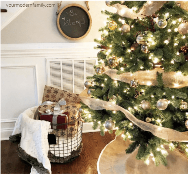 A close up of a Christmas tree with a basket of blankets and pillows beside it.