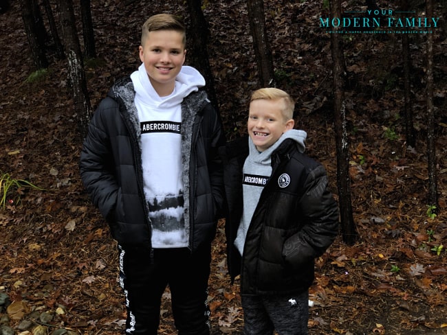 A couple of young boys standing next to each other wearing winter coats with trees in the background.