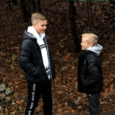 A couple of boys facing each other talking while standing in the woods.