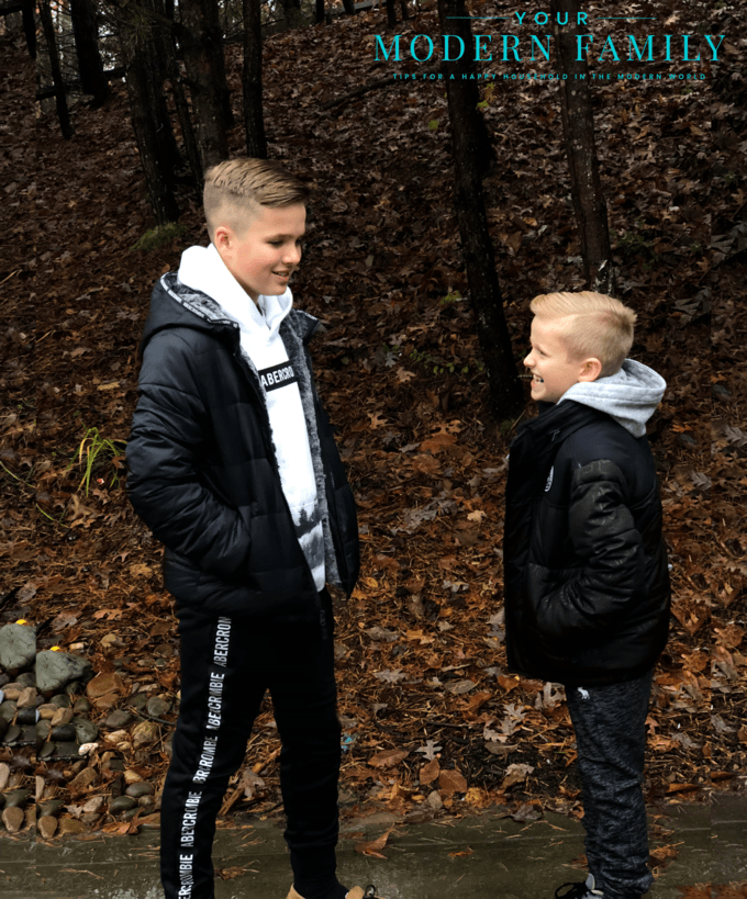 A couple of boys facing each other talking while standing in the woods.