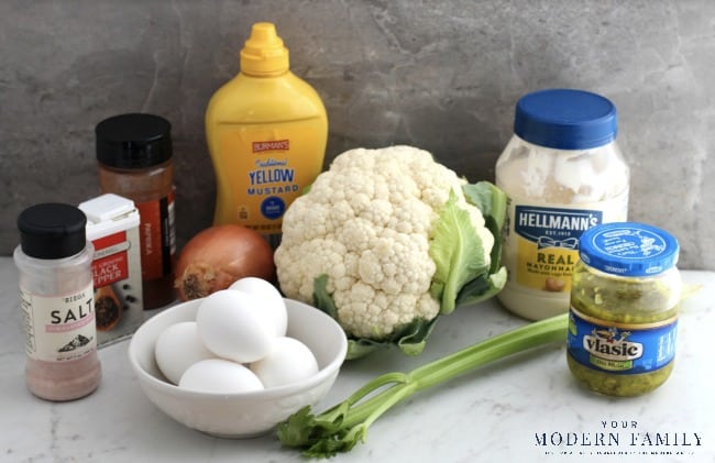 A bowl off eggs,  jars of mayonnaise and mustard, an onion and Cauliflower sitting on a table.