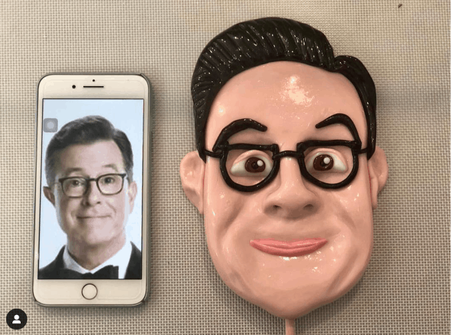 Stephen Colbert  posing for the camera on a cell phone with a giant look a like lollipop sitting on the table beside him.