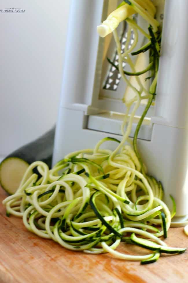 A spiralizer machine cutting zucchini into a pile of noodles on the table.