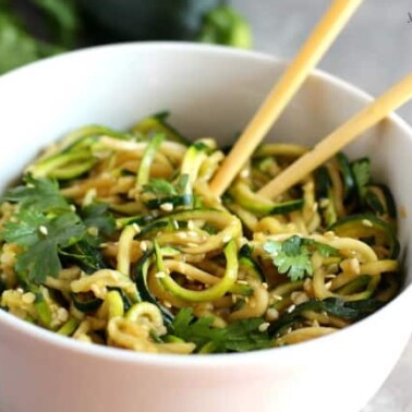 A white bowl with raw zucchini noodles with seasoning and sauce on them with chopsticks.