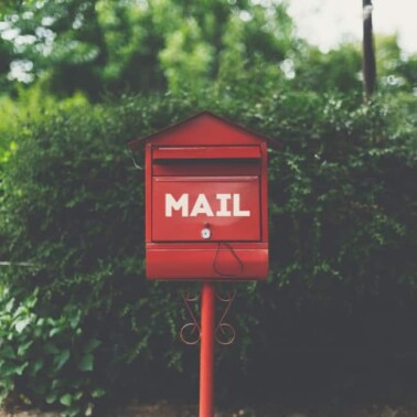 A red mailbox on a red pole with shrubs behind it.