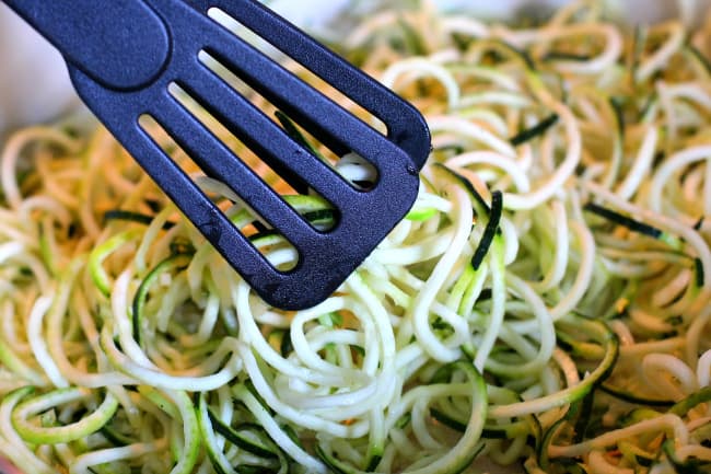 Raw zucchini noodles in a white bowl with black plastic thongs picking up noodles.