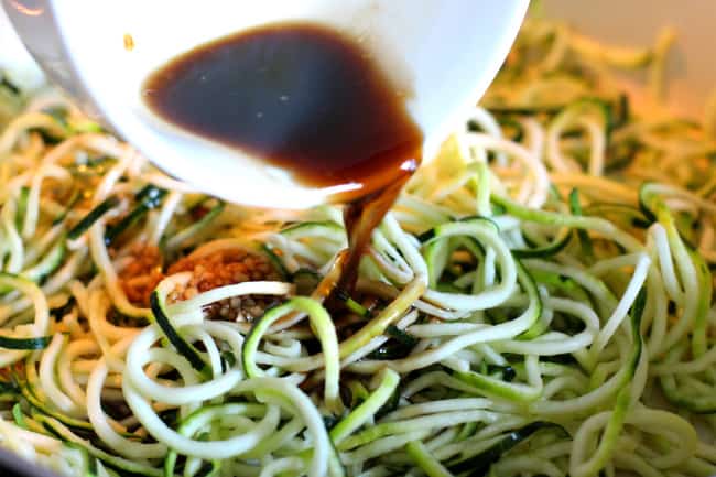 A white ladle pouring a dark colored sauce over top of zucchini noodles.