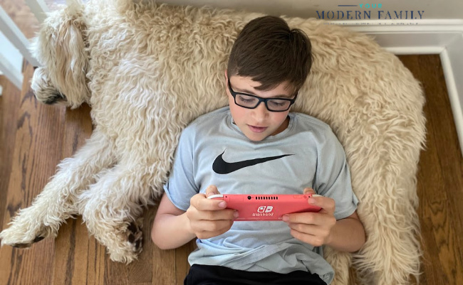 A boy lounging a dog while playing with a tablet.